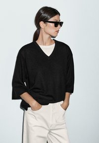 Black short-sleeve V-neck sweater over a light beige crew-neck top, paired with high-waisted white pants. Smooth texture, relaxed fit.