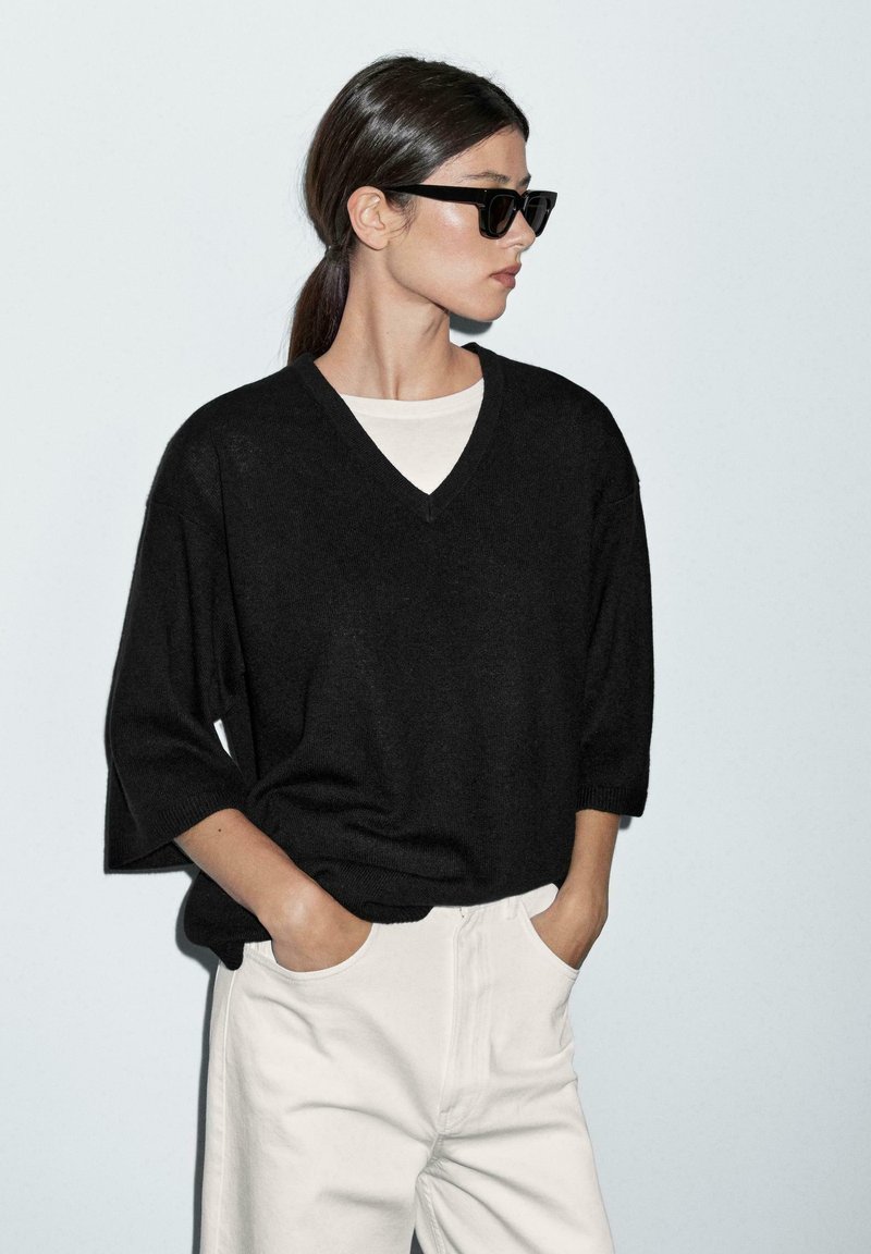 Black short-sleeve V-neck sweater over a light beige crew-neck top, paired with high-waisted white pants. Smooth texture, relaxed fit.