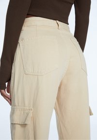 Kobieta ubrana w beżowe spodnie cargo z dużymi tylnymi i bocznymi kieszeniami, w połączeniu z dopasowanym brązowym swetrem z długim rękawem, ręka widoczna z boku.