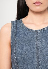 Gros plan d'une personne portant un haut en denim bleu sans manches, montrant le décolleté, l'épaule et une partie des longs cheveux foncés.