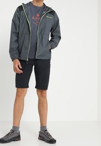 Herrarnas lätta huva-jacka i mörkgrått med limegröna detaljer, i kombination med svarta shorts och grå, slitstarka vandringsskor.