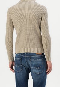 Persona che indossa un maglione a maglia beige testurizzato e jeans di denim blu, vista da dietro su uno sfondo bianco semplice.