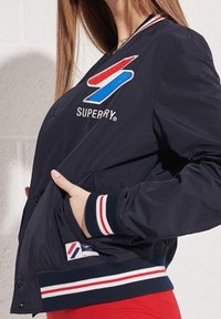 Marinblå bomberjacka med broderad logotyp, röda och blå accenter, vita randiga ribbade manschetter och sidofickor. Texturen verkar slät och lättvikts.