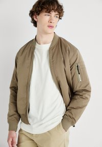Jachetă bomber maro cu fermoar frontala, manșete ajustate și un buzunar cu fermoar pe mâneca stângă. Pur tată peste o cămașă albă, evidențiind un look casual.
