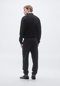 Sweatshirt preta e calças de treino combinando, feitas de tecido macio, com riscas laterais, e ténis pretos com solas brancas e detalhes às riscas.