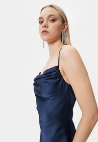 Navy Satin-Kleid mit einem Cowl-Ausschnitt und schmalen Trägern, das eine glatte Textur und eine figurbetonte Silhouette aufweist, ergänzt durch lange silberne Ohrringe.