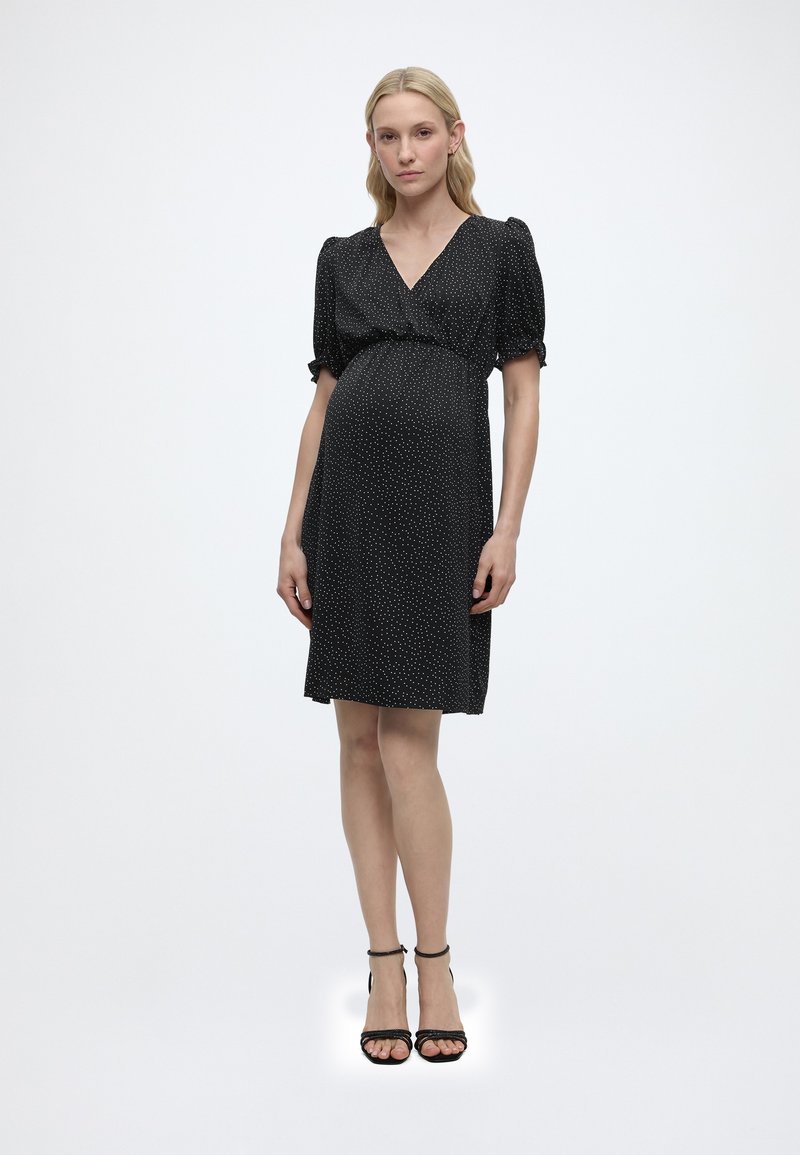 Schwangere Frau steht, trägt ein schwarzes knielanges Kleid mit weißen Punkten und schwarze Absatzschuhe mit Knöchelriemen, vor einem schlichten weißen Hintergrund.