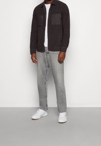 Giacca button-up grigio scuro con due tasche sul petto, abbinata a jeans grigio chiaro e sneakers bianche. Design semplice con una vestibilità rilassata.