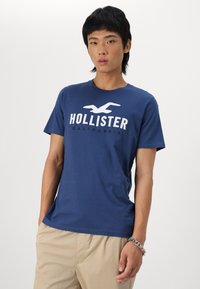 Granatowa bawełniana koszulka z okrągłym dekoltem, z białą grafiką "HOLLISTER CALIFORNIA" na klatce piersiowej. Krótkie rękawy, gładka tekstura.