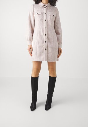 Kobieta w jasnoróżowej krótkiej sukience na guziki z długimi rękawami i kieszeniami z patkami, nosząca czarne kozaki do kolan.