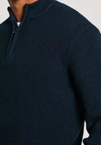 Marineblauer Strickpullover mit hohem Kragen und kurzem Reißverschluss, der ein strukturiertes, waffelartiges Muster aufweist. Die Nähte sind markant.