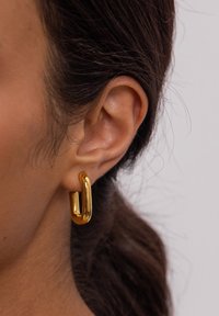 Goldene Creole mit U-förmigem Design, polierter Oberfläche und kräftigem Erscheinungsbild, getragen am menschlichen Ohr, das einen reichen metallischen Glanz zeigt.