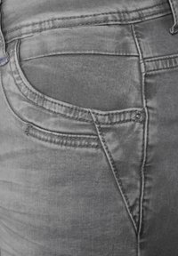 Šedé džíny z denimu s zadní kapsou, která má design zigzag stehu a kovový knoflík poblíž okraje kapsy. Hladká, texturovaná tkanina.