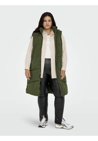 Chaleco acolchado verde con cuello alto, camisa blanca abotonada debajo, pantalones ajustados negros y zapatillas deportivas. Presenta un cierre de cremallera y bolsillos laterales.