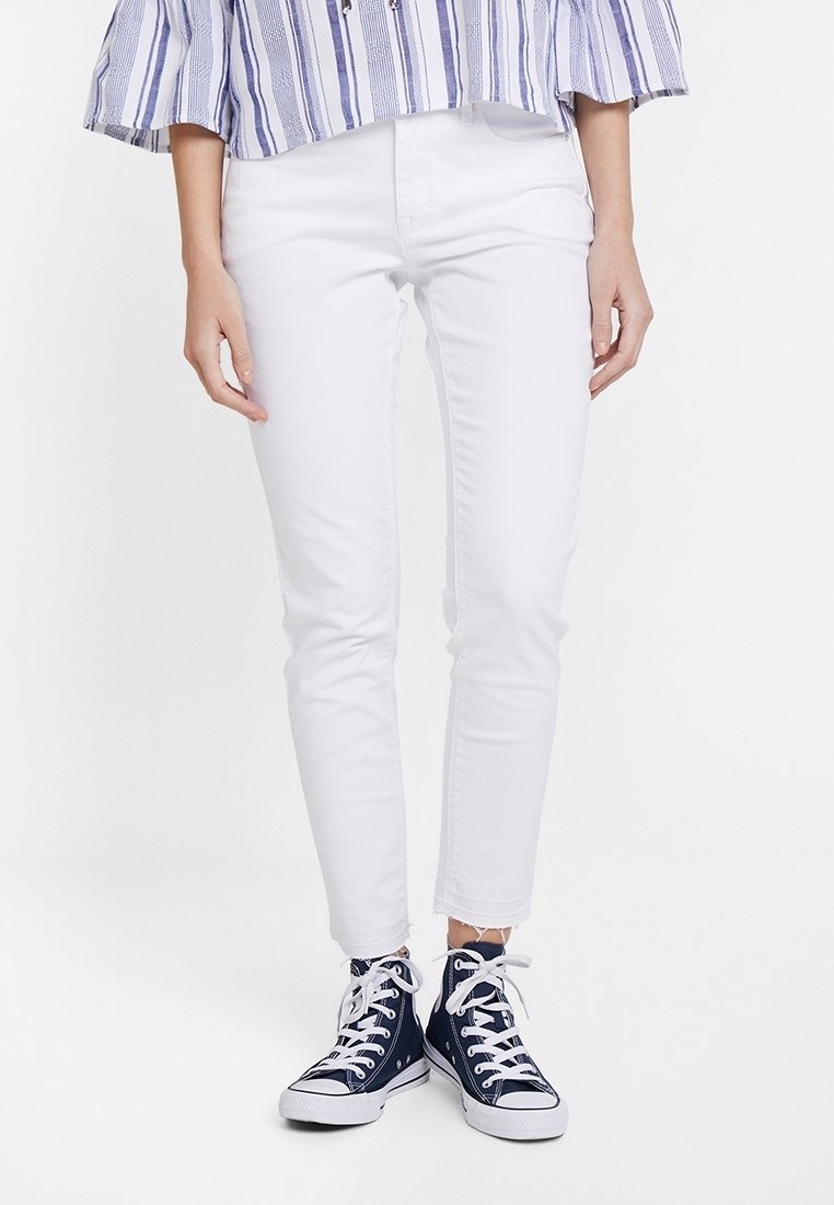 Vita slim-fit jeans med en lätt slitning vid fållen, parat med navy hög-top sneakers med vita snören och prickiga detaljer.