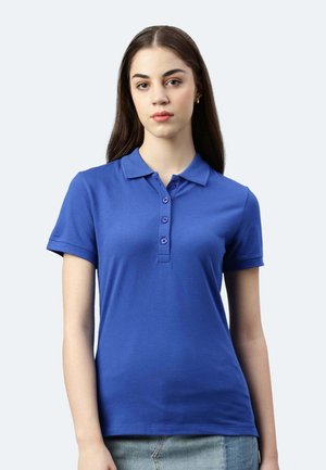 Jeune femme aux longs cheveux foncés portant un polo bleu à manches courtes et une jupe en denim clair, debout devant un fond clair.