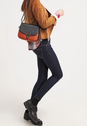 Brun mockaskinnjacka med vit sömnad, marinblå tajta jeans, svarta skor och en tvåfärgad axelväska med svart lock och orange kropp.