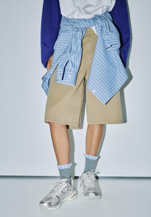 Osoba nosiaca béžové šortky po kolená, modrú pruhovanú košeľu zviazanú okolo pása, sivé volánikové ponožky a strieborné tenisky, stojaca pri bielej stene.