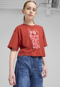 Rdeča skrajšana majica iz bombaža s kratkimi rokavi, ki prikazuje roza risanega sova grafiko in napis "Puma Sport Girls Club." Nosijo jo z modrimi jeans hlačami.
