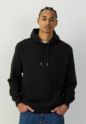 Young man with braided hair wearing a black hoodie and blue jeans, standing with hands in pockets against a plain background.