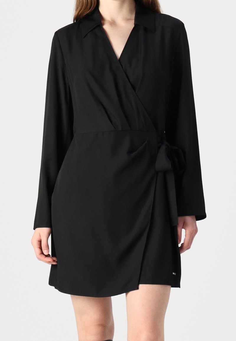 Femme portant une robe portefeuille noire à manches longues avec un lien sur le côté et un col, vue de la mi-cuisse au cou sur un fond clair.
