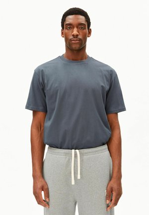 Mann trägt ein schlichtes dunkelgraues T-Shirt mit Rundhalsausschnitt und hellgraue Jogginghose mit weißem Kordelzug, steht vor weißem Hintergrund.