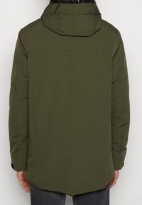 Giacca imbottita verde oliva con cappuccio, caratterizzata da una texture liscia e un orlo posteriore leggermente più lungo. Include polsini elasticizzati e dettagli di design minimali.