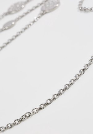 Chaîne métallique argentée avec des maillons ronds sur un fond clair, présentant des détails flous de bijoux en argent en arrière-plan supérieur.