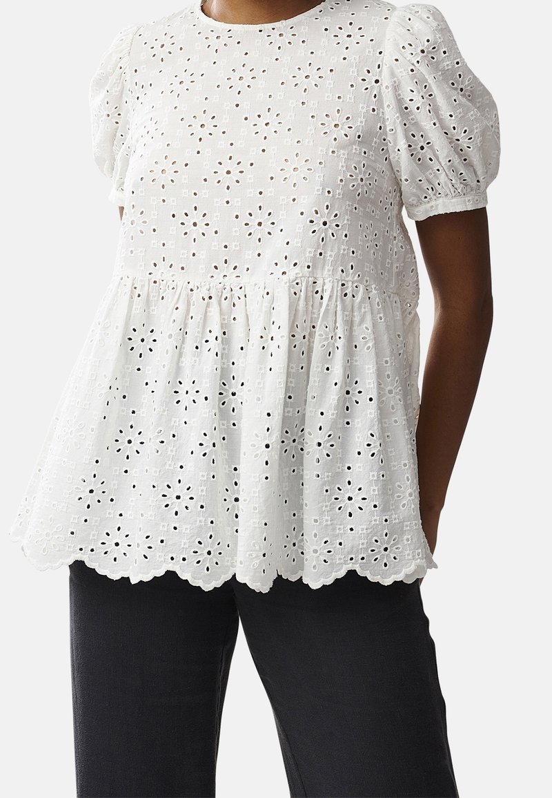 Blouse en dentelle ajourée blanche avec manches bouffantes et ourlet festonné portée avec un pantalon noir.