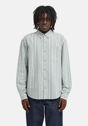 Adulte à la peau foncée et dreadlocks portant une chemise à rayures vert clair et blanche boutonnée et un jean bleu foncé, faisant face à l'avant.