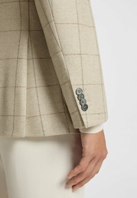 Beige rutigt kavaj i texturerat tyg, med fyra mörka knappar på ärmen och en ribbad stickad innerärm.