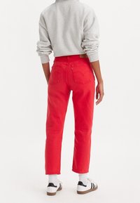 Jean en denim rouge à taille haute, jambes fuselées et deux poches arrière. Porté avec un sweat-shirt gris et des baskets blanches avec des accents noirs.