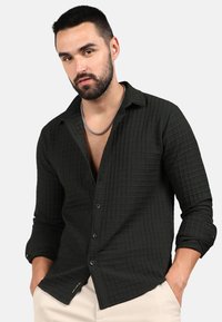 Camicia a quadri verde scuro in un tessuto testurizzato, con colletto, chiusura frontale con bottoni e maniche arrotolate. Dettaglio in catena argentata.