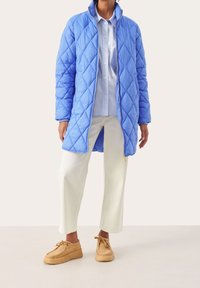 Jasnoniebieska pikowana kurtka puchowa z wysokim kołnierzem, z zapięciem na zamek, zestawiona z jasnoniebieską koszulą i białymi spodniami w szerokim kroju. Beżowe buty.