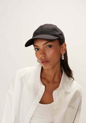 Casquette noire avec un visière courbée, arborant un petit logo bleu à l'avant. Le modèle porte une chemise blanche et un débardeur à la texture lisse.