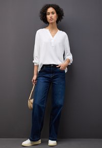 Blusa bianca a maniche lunghe con scollo a V, abbinata a jeans a gamba larga blu scuro. La modella tiene in mano una piccola borsa rotonda di paglia e indossa scarpe bianche.