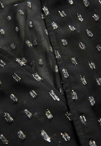 Tissu noir présentant un motif texturé de petites formes métalliques argentées, créant un contraste dimensionnel sur la surface.