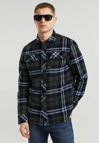 Groen en marineblauw geruit flanellen shirt met twee borstzakken, knoopsluiting en een gestructureerde stof. Draagt met donkere zonnebrillen en jeans.