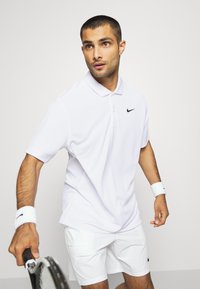 Balta Nike polo marškinėliai su juodu logotipu, trumpi rankoviai ir tradicinė apykaklė. Derinami su baltais šortais ir balta apyranke.