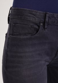 Jeans de mezclilla oscura con una textura suave, cuatro bolsillos y una cinturilla estándar con cierre de botón. Detalles de costura sutiles visibles.
