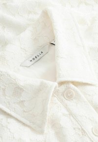 Weiße Spitzenbluse mit einem Kragen, zarten Blumenmustern und einer geknöpften Vorderseite, ausgestattet mit einem weichen Innenfutter und einem Markenetikett.