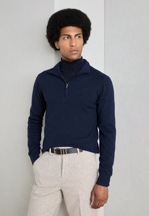 Navy pullover sweater with a zippered collar, paired with light beige textured trousers and a black belt, set against a neutral background.