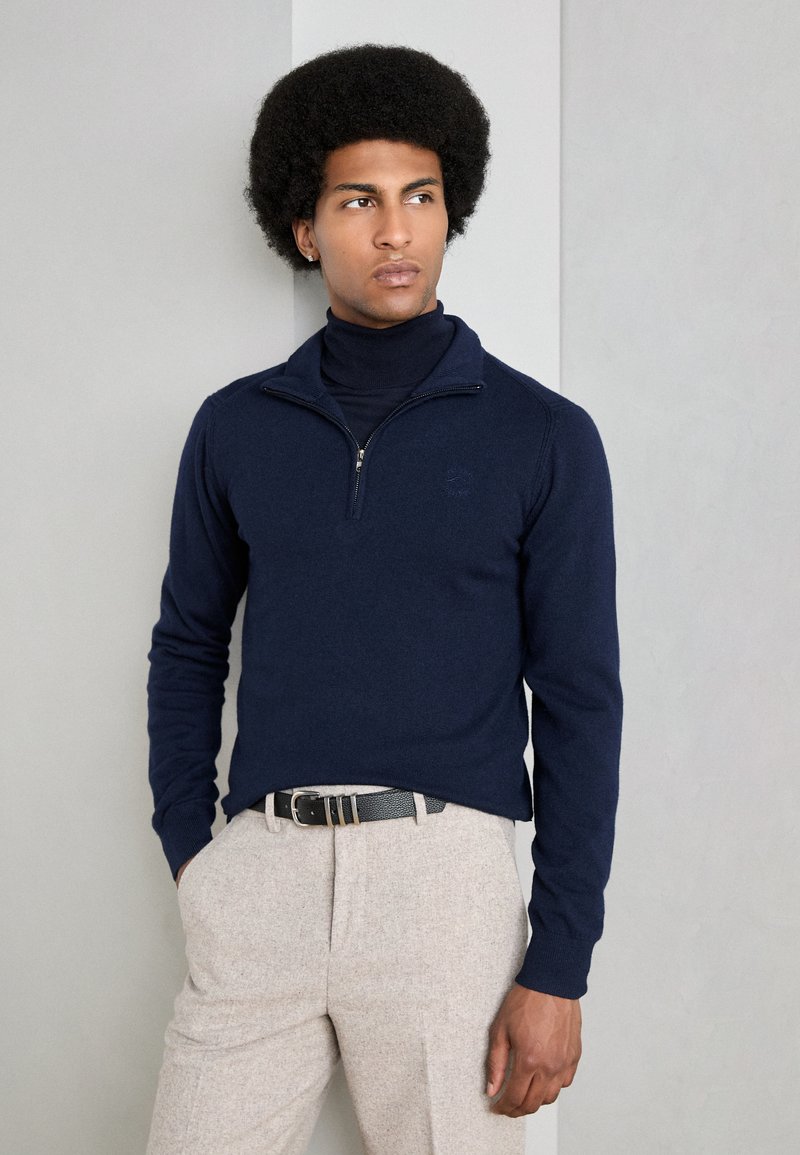 Suéter azul marino de cuello con cremallera, combinado con pantalones beige claros texturizados y un cinturón negro, sobre un fondo neutro.