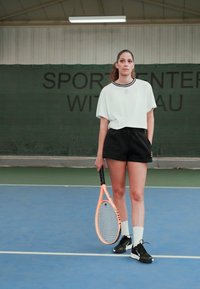 Camisa branca de mangas curtas com detalhes pretos, calções pretos e sapatilhas desportivas pretas. Segurando uma raquete de ténis laranja num court azul.