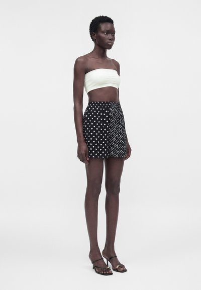 Ψηλή γυναίκα που φοράει λευκό στράπλες crop top, μαύρη μίνι φούστα με σχέδια αστεριών και κυμάτων, και μαύρα ανοιχτά σανδάλια με τακούνια.