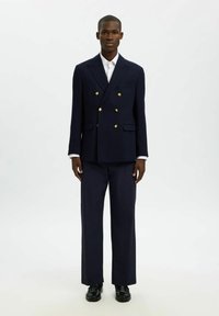 Granatowy dwurzędowy blezer z złotymi guzikami, dopasowane spodnie, biała koszula i czarne buty. Krój dopasowany, gładka faktura.