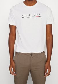 Muž v bílém tričku Hilfiger New York a hnědých kalhotách na knoflíky na jednobarevném pozadí.