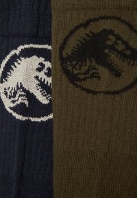 Dois pares de meias: um par em azul-marinho com um gráfico de dinossauro branco, e o outro em verde-azeitona com um gráfico de dinossauro preto. Tecido de malha texturizada.