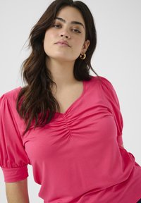 Haut rose à manches courtes avec un décolleté en V et un détail froncé au niveau de la poitrine. Texture douce et coupe décontractée. Comprend des manches bouffantes.
