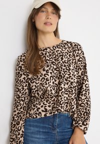 Blusa de estampado de leopardo en beige y marrón, con un escote redondo, detalles plisados y mangas largas. Combinada con jeans de mezclilla azul.